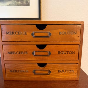 May include: A wooden storage chest with three horizontal drawers. Each drawer features a dark metal label holder and the black printed text "MERCERIE" on the left and "BOUTON" on the right. The wood has a medium brown finish with visible grain.