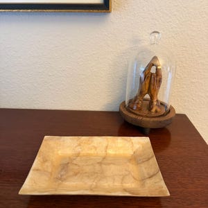 Könnte beinhalten: Ein rechteckiges Tablett mit hellbeiger, marmorierter Oberfläche. Dahinter befindet sich eine Holzskulptur von zwei gefalteten Händen, die unter einer gläsernen Kuppel auf einem runden Holzsockel platziert ist.