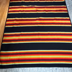 May include: A rectangular blanket featuring a repeating pattern of wide black horizontal bands alternating with vibrant, multi-colored striped sections in shades of red, orange, yellow, and green, creating a bold, high-contrast design.