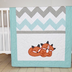 May include: A white and turquoise chevron baby quilt with a border of turquoise and a design of three orange foxes sleeping on a white background.