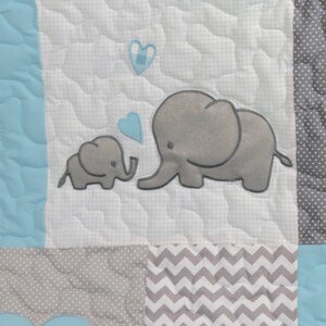 May include: A white baby quilt with a gray and white chevron pattern, a gray and white polka dot pattern, and a light blue solid pattern. The quilt features two gray embroidered elephants with blue hearts.