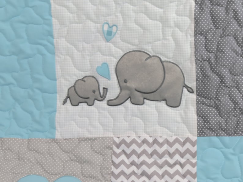 May include: A white baby quilt with a gray and white chevron pattern, a gray and white polka dot pattern, and a light blue solid pattern. The quilt features two gray embroidered elephants with blue hearts.