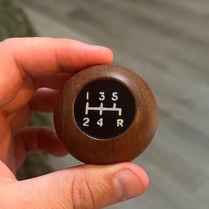 May include: A spherical wooden gear shift knob featuring a dark brown wood grain finish. The top surface has a circular black inlay displaying a white manual transmission shift pattern with numbers 1, 2, 3, 4, 5 and the letter R in yellow.