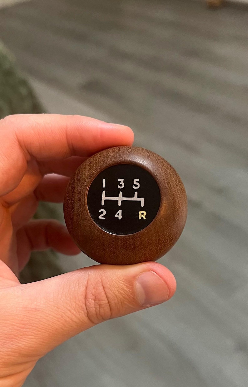 May include: A spherical wooden gear shift knob featuring a dark brown wood grain finish. The top surface has a circular black inlay displaying a white manual transmission shift pattern with numbers 1, 2, 3, 4, 5 and the letter R in yellow.