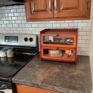 May include: A two-tier wooden bread box with a dark reddish-brown finish and clear glass front panels. Each tier features a small black knob for opening the drop-down doors, providing a visible storage space for loaves of bread on a kitchen counter.