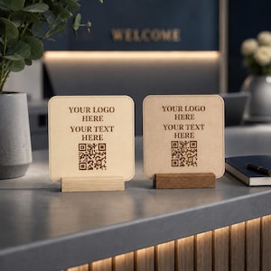 May include: Two square light-colored wooden signs with rounded corners, each held upright by a rectangular wooden base. The signs feature the engraved text "YOUR LOGO HERE" and "YOUR TEXT HERE" above a square QR code in a dark brown finish.