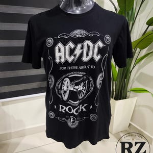 May include: A black short-sleeve t-shirt featuring a white graphic print of the AC/DC logo, the text "FOR THOSE ABOUT TO ROCK," and an illustration of a cannon. The design includes decorative scrollwork borders and circular accents on the front.