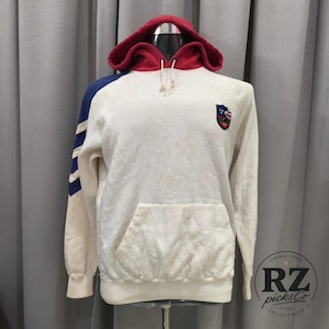 Puede incluir: Una sudadera con capucha de color crema y manga larga que presenta una capucha roja con cordones blancos, un panel azul en la manga con rayas blancas y un pequeño parche con escudo en el pecho izquierdo. Incluye bolsillo tipo canguro.