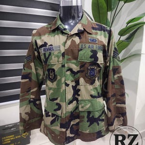 May include: A long-sleeve military camouflage jacket featuring a woodland pattern of green, brown, and black. The jacket includes a U.S. AIR FORCE patch, a name tag reading MEMMEL, two chest pockets, and various embroidered military insignia patches.