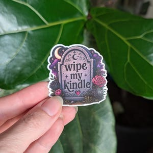 May include: A die-cut sticker featuring a purple and black tombstone illustration with the text "wipe my kindle" in the center. The design includes a crescent moon, stars, and red roses surrounding the base of the tombstone with a small heart.