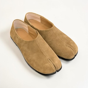 May include: A pair of tan suede split-toe shoes featuring a low-profile design with a central seam running down the vamp. The interior is lined with a light beige material, and the shoes are set on a thin black sole, showcasing a distinct toe separation.