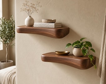 May include: Two curved, solid dark wood floating shelves mounted on a wall. The top shelf holds a white vase with dried flowers and two stacked books. The bottom shelf features a small gold bowl and a potted green plant with trailing leaves.