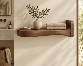 May include: A wall-mounted dark wood floating shelf with a curved edge and a circular recessed cutout. The shelf holds a small stone bowl, a round beige vase containing green olive branches, and a stack of two folded beige towels.