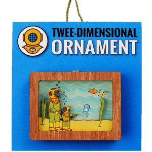 May include: A wooden ornament depicting two divers in a blue and green underwater scene. The ornament is framed in wood and has a string for hanging. The text "TWEEDIMENSIONAL ORNAMENT" is printed on the card behind the ornament.