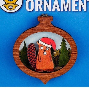 May include: A wooden Christmas ornament shaped like a bauble with a beaver wearing a Santa hat inside. The ornament has a brown wood finish and a blue background with the text "Twee-Dimensional Ornament".