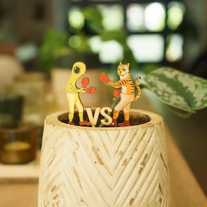 May include: A white ceramic planter with a chevron pattern holds two wooden figurines. One figurine is a yellow humanoid with red boxing gloves, the other is a tiger with orange and black stripes wearing red boxing gloves. The figurines are facing each other and the word "VS" is in between them.