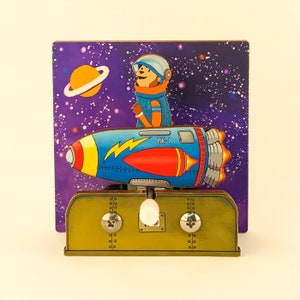 May include: A blue and yellow rocket ship with a red nose cone and the text "MK-1" on the side. A cartoon bear wearing a blue spacesuit is standing in the rocket. The rocket is on a wooden base with a starry purple background.