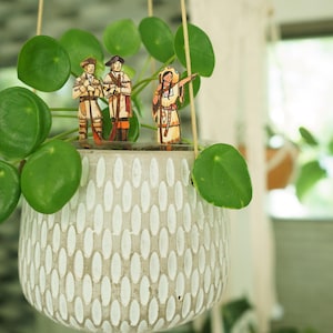 May include: A hanging planter with a white speckled finish, filled with green leafy plants. Three small figurines of people stand in the planter, two men and one woman.