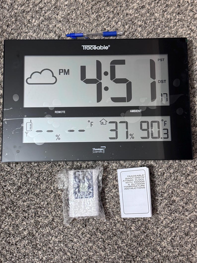 May include: A black Traceable digital atomic clock with a large display showing 4:51 PM, a cloud icon, and temperature readings of 37% humidity and 90.3 degrees Fahrenheit. Includes a blue pen, a remote sensor in bubble wrap, and an instruction manual.