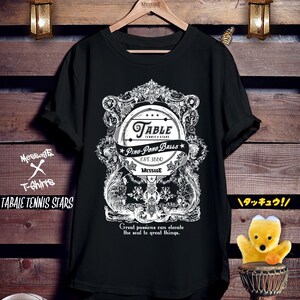 以下が含まれることがあります： 黒いTシャツで、装飾的なフレームの中に卓球のラケットの白いグラフィックが描かれています。テキストには「Table Tennis Stars」、「Ping-Pong Balls」、「Est. 1880」、「Message」、「Great passions can elevate the soul to great things」と書かれています。