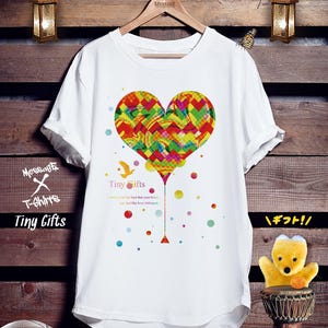 Peut inclure: Un t-shirt blanc à manches courtes orné d'un grand cœur multicolore au motif chevron et marbré. Sous le cœur, le texte Tiny Gifts est inscrit avec la silhouette d'un petit oiseau et des points colorés dispersés.
