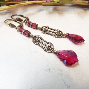 May include: A pair of dangle earrings with a vintage style design. The earrings feature a long, rectangular pendant with intricate details and a teardrop-shaped crystal. The crystals are a deep pink color.