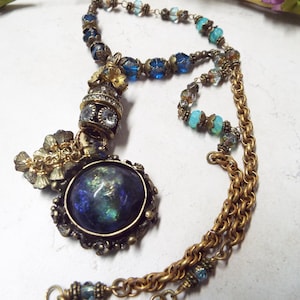 Puede incluir: Un collar de cadena dorada con un colgante de piedra iridiscente azul y verde. El colgante está rodeado de filigrana dorada y tiene un pequeño amuleto dorado adjunto. La cadena está hecha de pequeñas cuentas doradas y tiene un acento de cuentas de vidrio azul y verde.