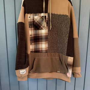 May include: A long-sleeve hooded sweatshirt featuring a patchwork design in various shades of brown, tan, and cream. The garment includes a plaid chest pocket, a front kangaroo pocket, a drawstring hood, and a small embroidered bear face on the cuff.