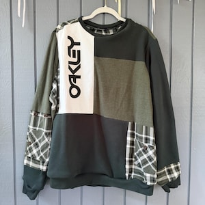 May include: A long-sleeve crewneck sweatshirt featuring a patchwork design of solid dark green fabric and green, white, and black plaid panels. The front includes a vertical white rectangular panel with the black text OAKLEY printed on it.