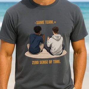 May include: A charcoal gray crew neck t-shirt featuring a graphic of two people sitting on a round rug playing video games. The text above reads "SAME TEAM." and the text below reads "ZERO SENSE OF TIME." The design is centered on the chest.