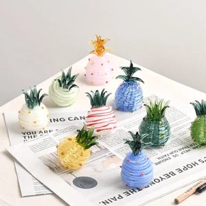 May include: A collection of nine small glass pineapple figurines in various colors including white, light green, pink, red-striped, blue, dark green, yellow, and light blue, each topped with a dark green glass leaf crown and small dots.