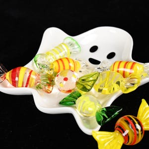 Pode incluir: Um prato de cerâmica branca em forma de fantasma contendo vários pequenos doces de vidro coloridos. Os doces de vidro apresentam vários padrões, incluindo listras amarelas e vermelhas, superfícies transparentes com pontos e detalhes verdes ou amarelos.
