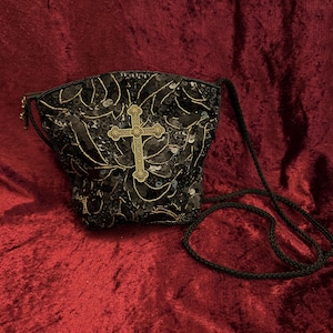 May include: Black sequined crossbody bag with a decorative gold cross. The bag has a black rope strap and a zip closure. The sequins create a textured surface, and the gold cross adds a striking contrast to the dark background.