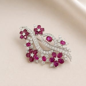 Può includere: Una spilla color argento caratterizzata da un design a nastro vorticoso incastonato con piccole pietre chiare. Il pezzo è decorato con molteplici grappoli floreali composti da pietre ovali rosa scuro e piccole pietre chiare al centro.