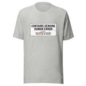 May include: A heather grey short-sleeved t-shirt featuring a rectangular graphic with the black text CONTAINS GENUINE HUMAN ERROR, the orange word WITH, and the text MADE WITHOUT ASSISTANCE RESULTS MAY BE SINCERE, plus a brown coffee ring stain.