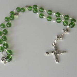 May include: A rosary with green glass beads and silver-toned metal accents. The beads are strung on a silver chain, and the cross features a woven design. The rosary is displayed on a white surface.