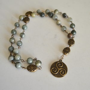May include: A necklace featuring round, gray-green beads strung on a delicate chain. The necklace includes ornate, bronze-colored connectors and a circular pendant with a swirling Celtic design.