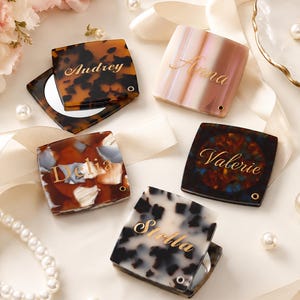 May include: Five square compact mirrors with hinged lids, each featuring a unique pattern and a gold-colored name engraved on the surface. The designs include brown tortoiseshell, pink and white stripes, multi-colored mosaic, dark tortoiseshell, and white with black spots.