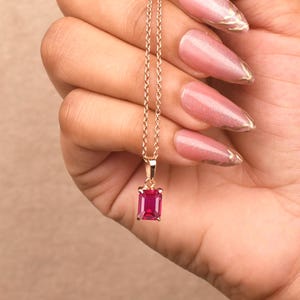 May include: A delicate rose gold chain necklace featuring a single rectangular emerald-cut vibrant pink gemstone pendant. The pendant is securely held by a small metal bail attached to the thin, polished rose gold link chain.