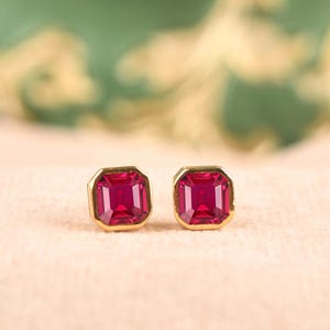 May include: A pair of stud earrings featuring vibrant, rectangular-cut red gemstones set in polished gold metal. The stones have a faceted surface that reflects light, and each is encased in a simple, clean gold bezel setting for a classic look.