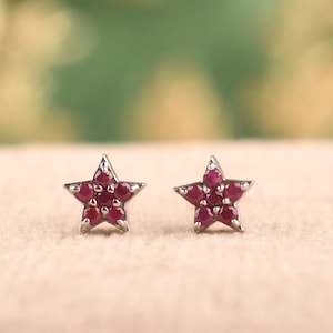 Puede incluir: Un par de pendientes de botón con forma de estrella, cada uno con cinco pequeñas piedras preciosas circulares de color rojo intenso engastadas en un marco metálico. El diseño de estrella es simétrico, con una piedra en cada punta.