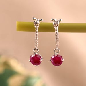 May include: A pair of silver hoop earrings featuring a thin band with small embedded stones and a single round, faceted deep red ruby-coloured gemstone dangling from the base of each hoop, suspended from a thin horizontal green rod.
