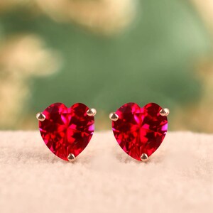 Puede incluir: Un par de pendientes de botón con gemas rojas facetadas en forma de corazón. Cada piedra está asegurada por pequeñas garras metálicas, creando un diseño claro y simétrico que resalta el color carmesí intenso y el corte preciso de las gemas.