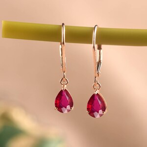 Peut inclure: Une paire de boucles d'oreilles en or rose avec fermoir levier, ornées de pierres précieuses rouge foncé en forme de poire à facettes. Les boucles d'oreilles sont suspendues à une fine tige verte horizontale, révélant l'éclat du métal.