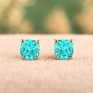 May include: A pair of stud earrings featuring square-cut, bright turquoise gemstones set in gold-toned metal prongs. The stones have a faceted surface that reflects light, and the earrings are positioned side by side on a soft, neutral-colored surface.