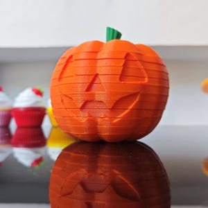 May include: A 3D printed orange pumpkin with a green stem and a carved jack-o'-lantern face featuring triangular eyes and a jagged mouth. The pumpkin has visible horizontal layer lines from the printing process and a smooth, rounded shape.