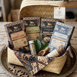 Puede incluir: Una cesta de bambú trenzado que contiene cuatro cajas rectangulares de café indonesio WoCA etiquetadas como Arabica Gayo, Arabica Toraja, Arabica Luwak y Bali Kintamani. Incluye un pequeño frasco de vidrio con granos de café verdes, una pala de madera y hojas.