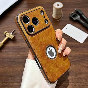 May include: A tan, textured faux leather phone case for an iPhone, featuring a prominent circular cutout in the center that reveals the Apple logo, a raised camera module housing three lenses, and black stitched edges along the perimeter.