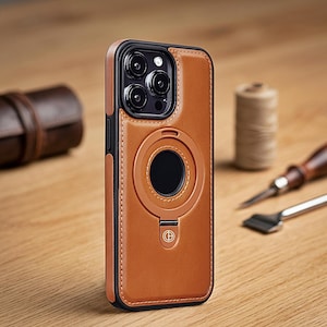 Könnte beinhalten: Eine Smartphone-Hülle aus hellbraunem Leder mit schwarzem Kamerarahmen und einem kreisförmigen magnetischen Ringständer auf der Rückseite. Die Hülle weist sichtbare Nähte an den Kanten und ein kleines kreisförmiges Logo im unteren Bereich auf. Das Design umfasst eine Aussparung für das Dreifach-Kameraobjektiv und einen schwarzen Rand um den Bildschirmbereich.
