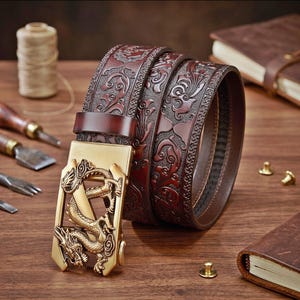 May include: A dark brown leather belt featuring an intricate embossed floral pattern and a gold-toned metal buckle shaped like a dragon. The belt is coiled, showcasing the detailed texture of the leather and the metallic finish of the buckle.